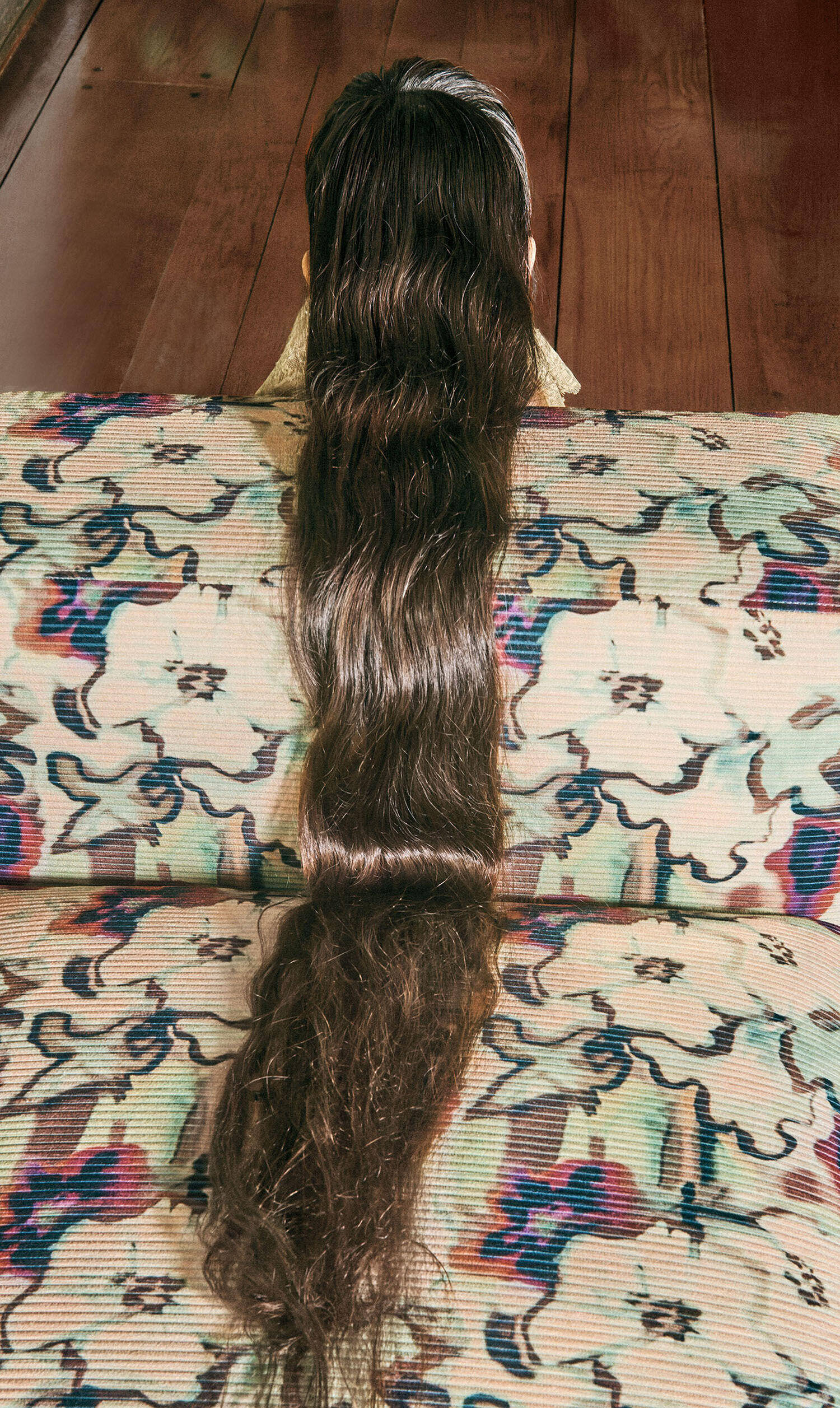A person seen from behind, with long, dark hair flowing down the back of a patterned, pleated armchair.