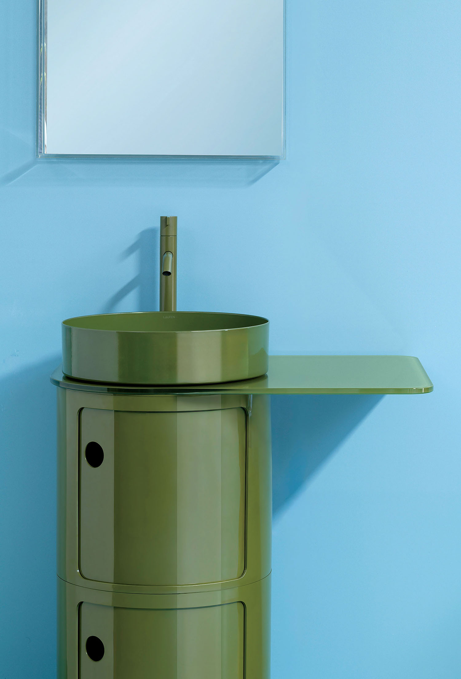 Olive green cylindrical bathroom vanity with a matching round vessel sink, tall faucet, and extended side shelf, mounted against a light blue wall with a rectangular mirror above.