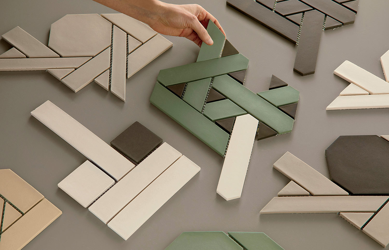 A hand arranges geometric ceramic tiles in green, brown, and neutral tones on a flat surface.