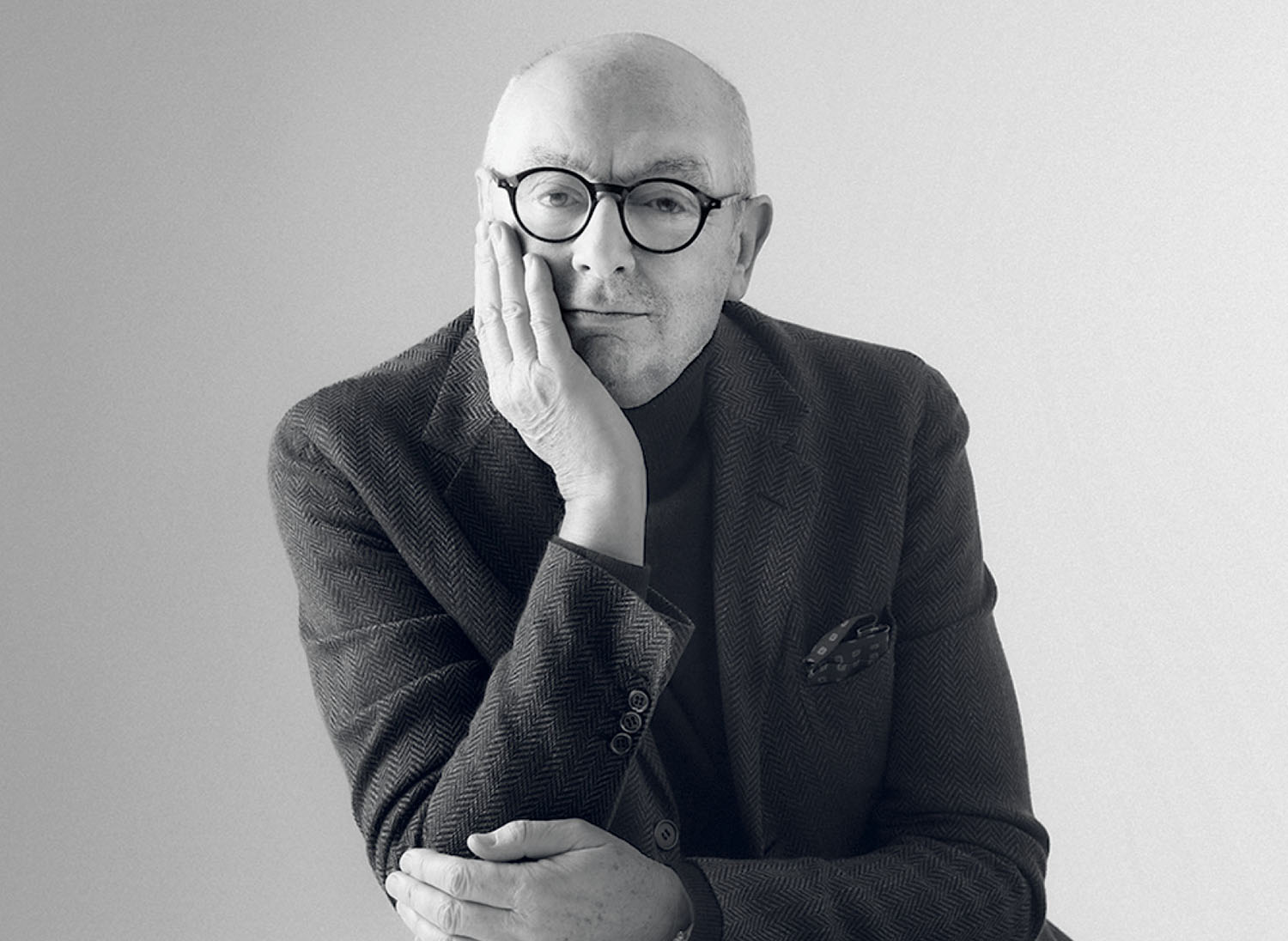 Portrait of a bald man in glasses and a blazer resting his cheek on his hand.