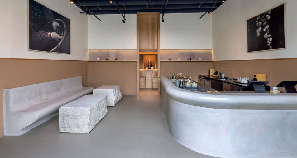 Interior of a minimalist cafe with curved concrete seating and counters, rough-hewn stone tables, and framed artwork on the walls.