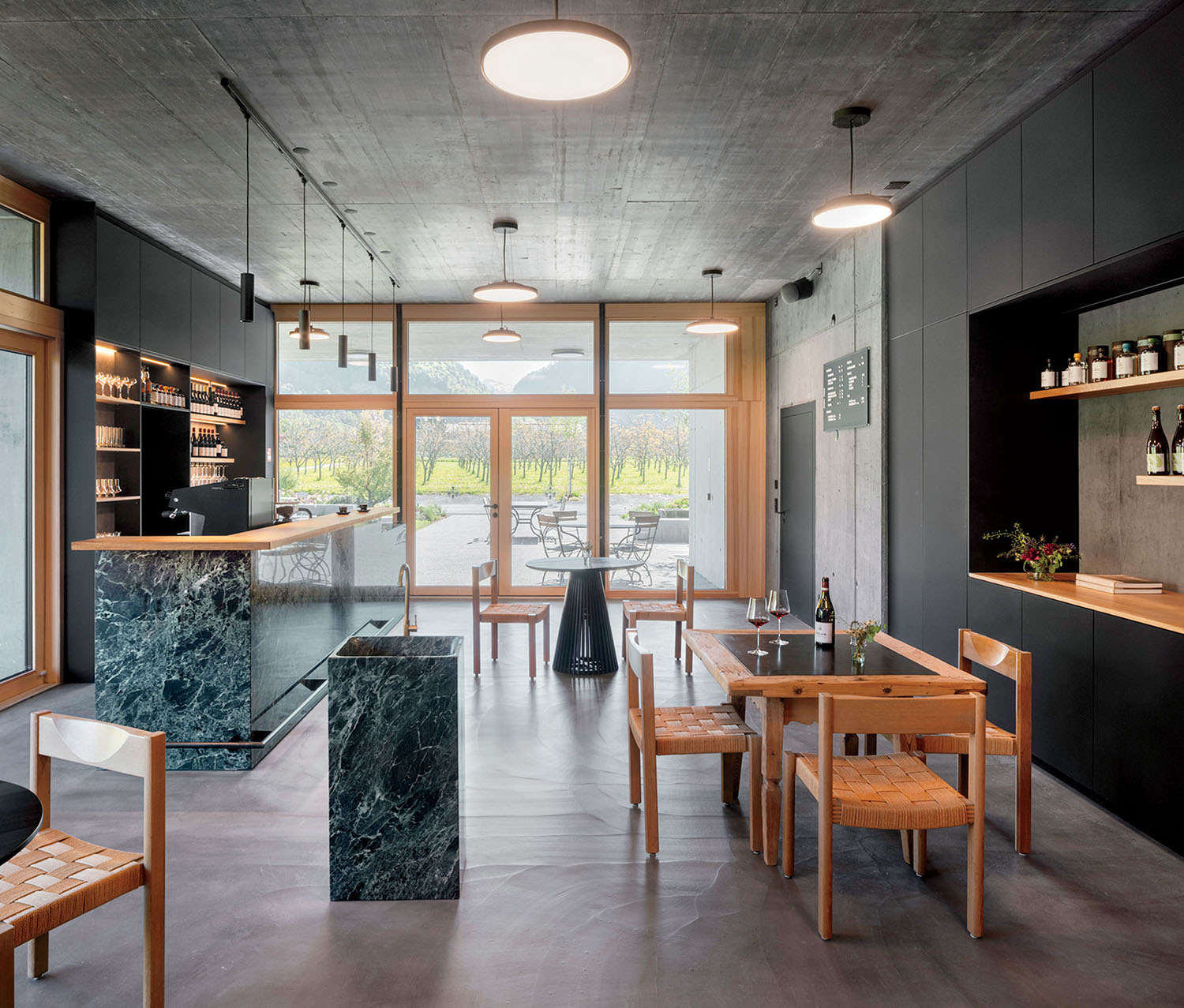 Interior of a modern tasting room with a dark green marble bar, wooden tables and chairs, and floor-to-ceiling windows looking out onto a vineyard.