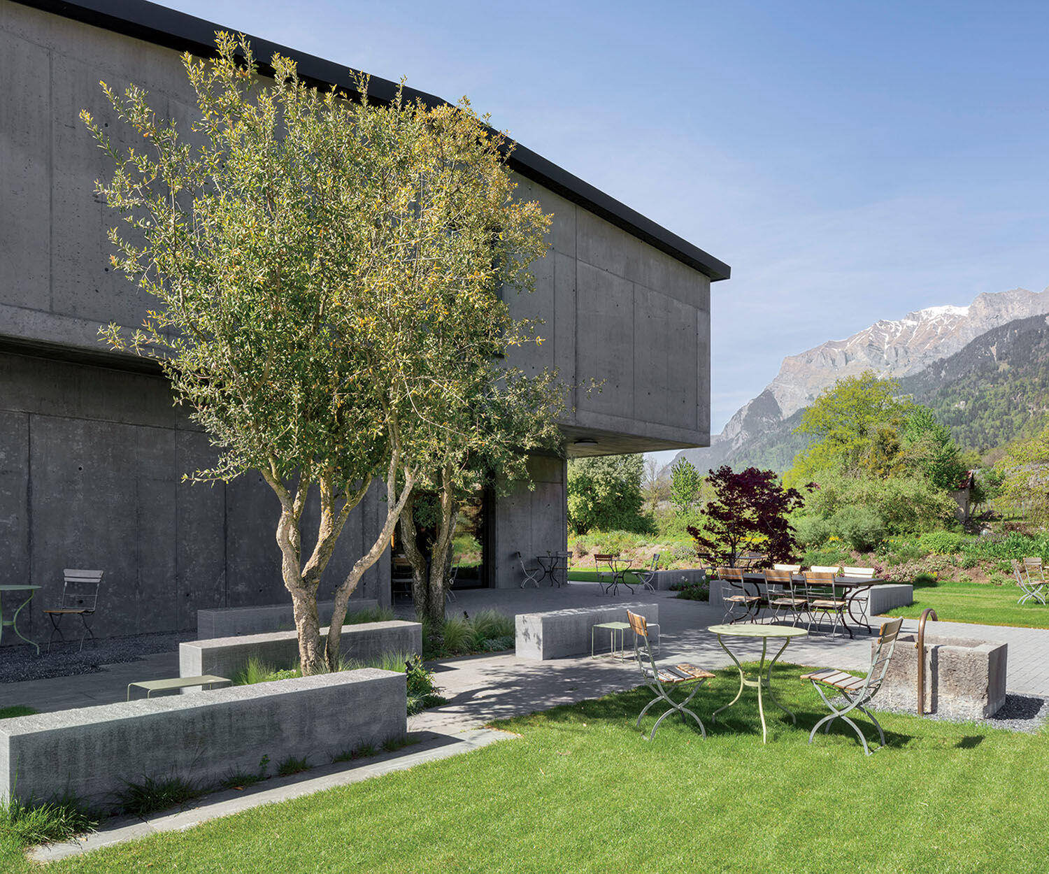 A modern concrete building with an outdoor patio featuring metal tables and chairs, surrounded by a lawn, trees, and mountain views.