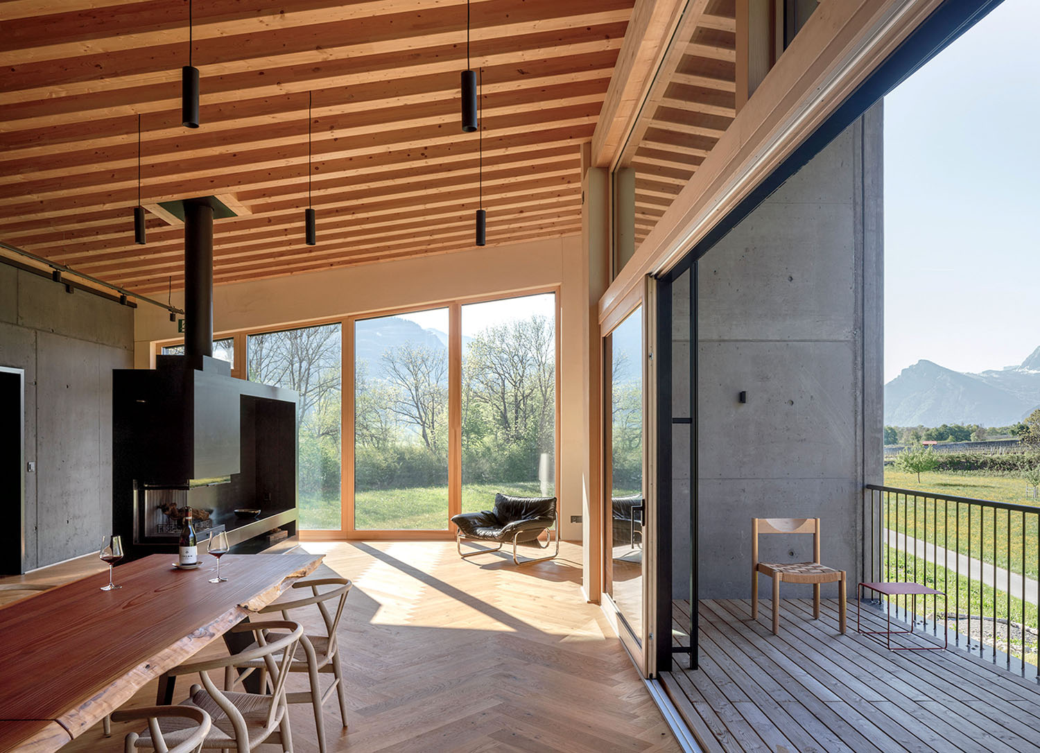 Modern dining area with a wooden table, fireplace, and floor-to-ceiling windows opening onto a balcony with a mountain view.