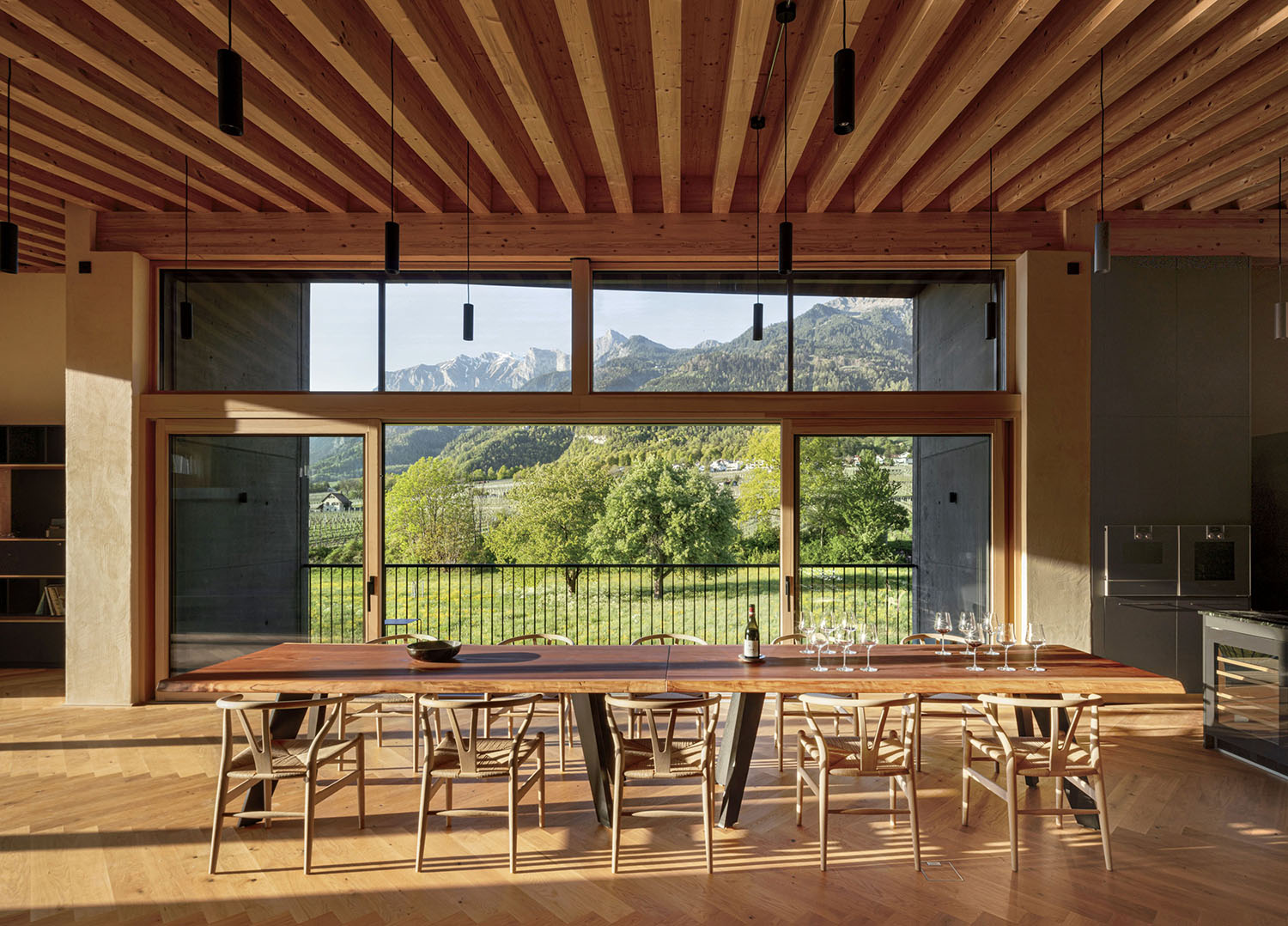 A long wooden dining table with eight chairs faces a large window overlooking a mountain landscape.
