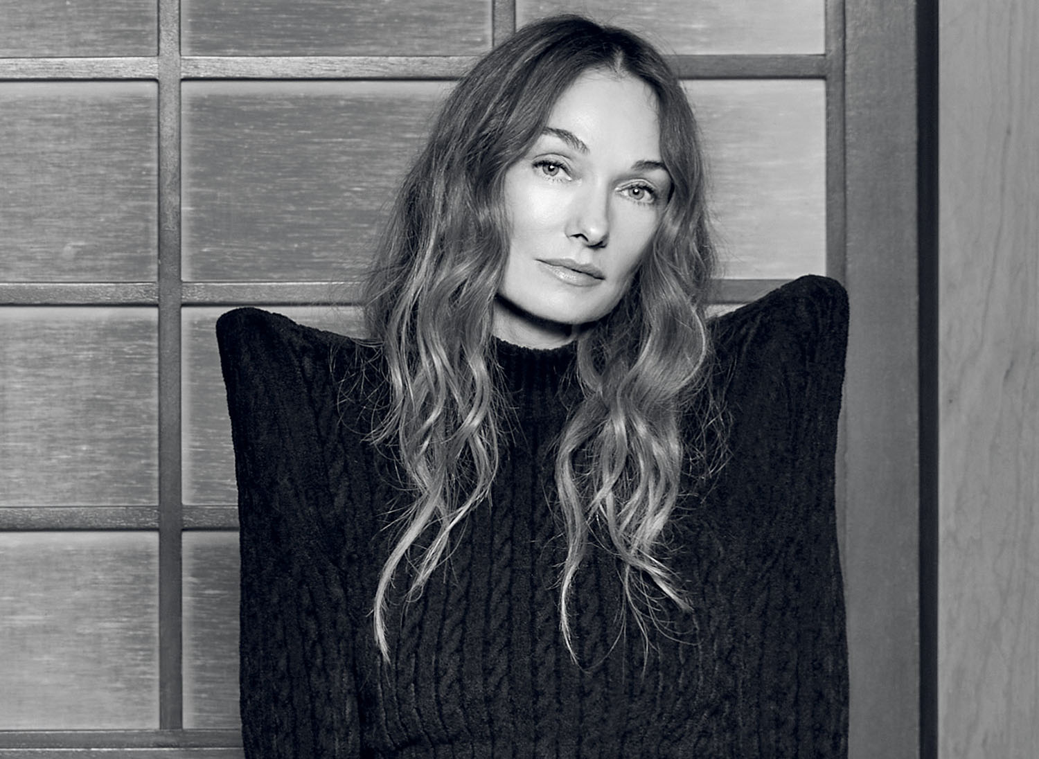 A black-and-white portrait of a woman with long, wavy hair wearing a high-neck sweater with prominent, structured shoulders.