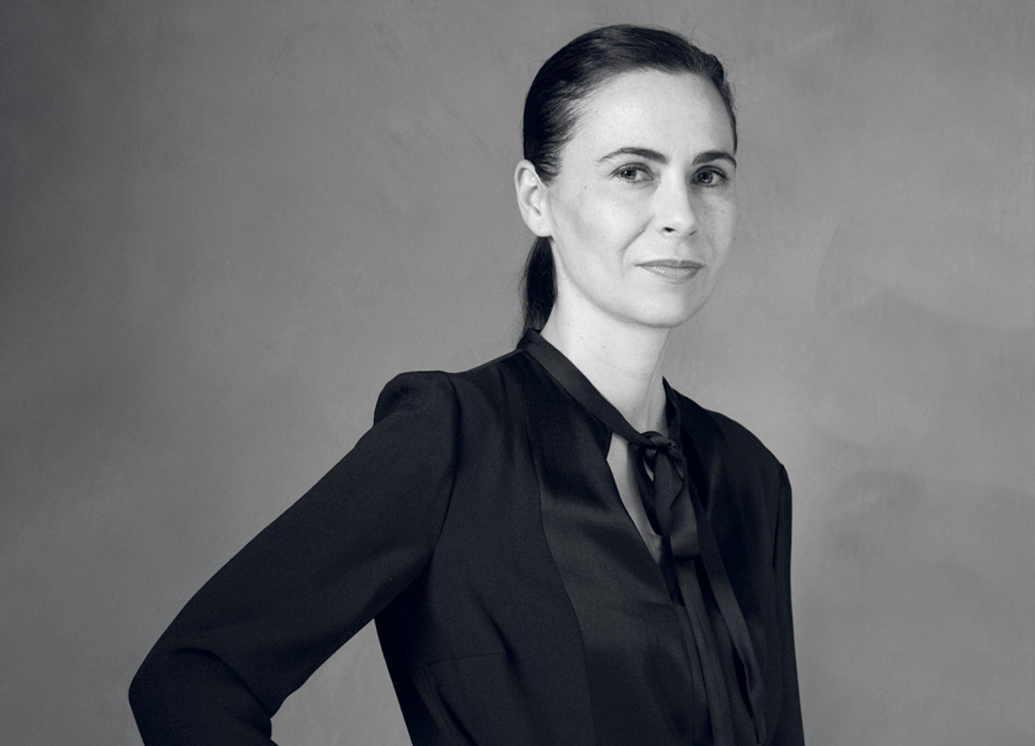 A woman with her hair pulled back, wearing a black collared blouse with a tie detail.