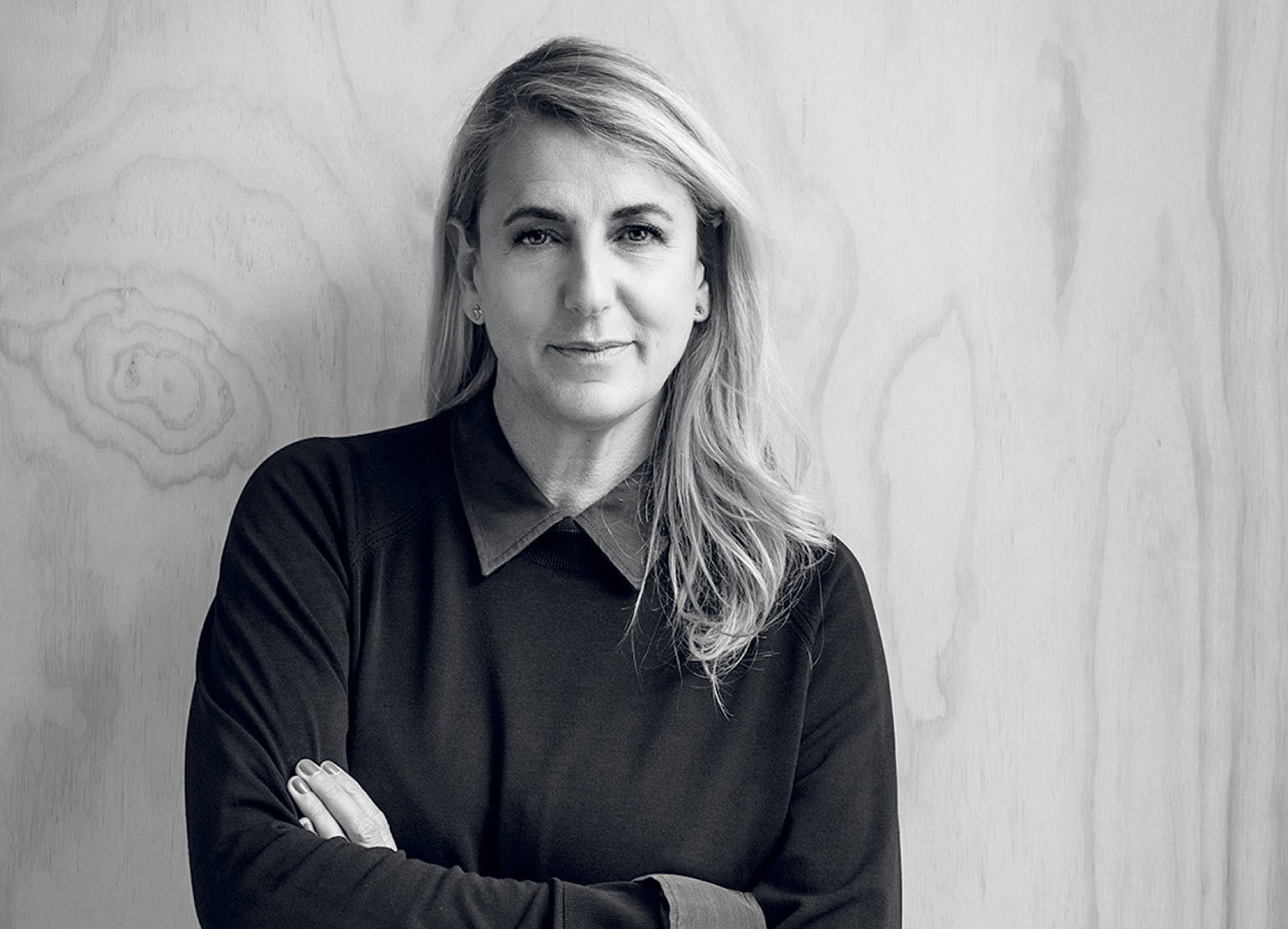 A woman with shoulder-length hair wearing a dark collared shirt, shown from the chest up against a wood-paneled wall.