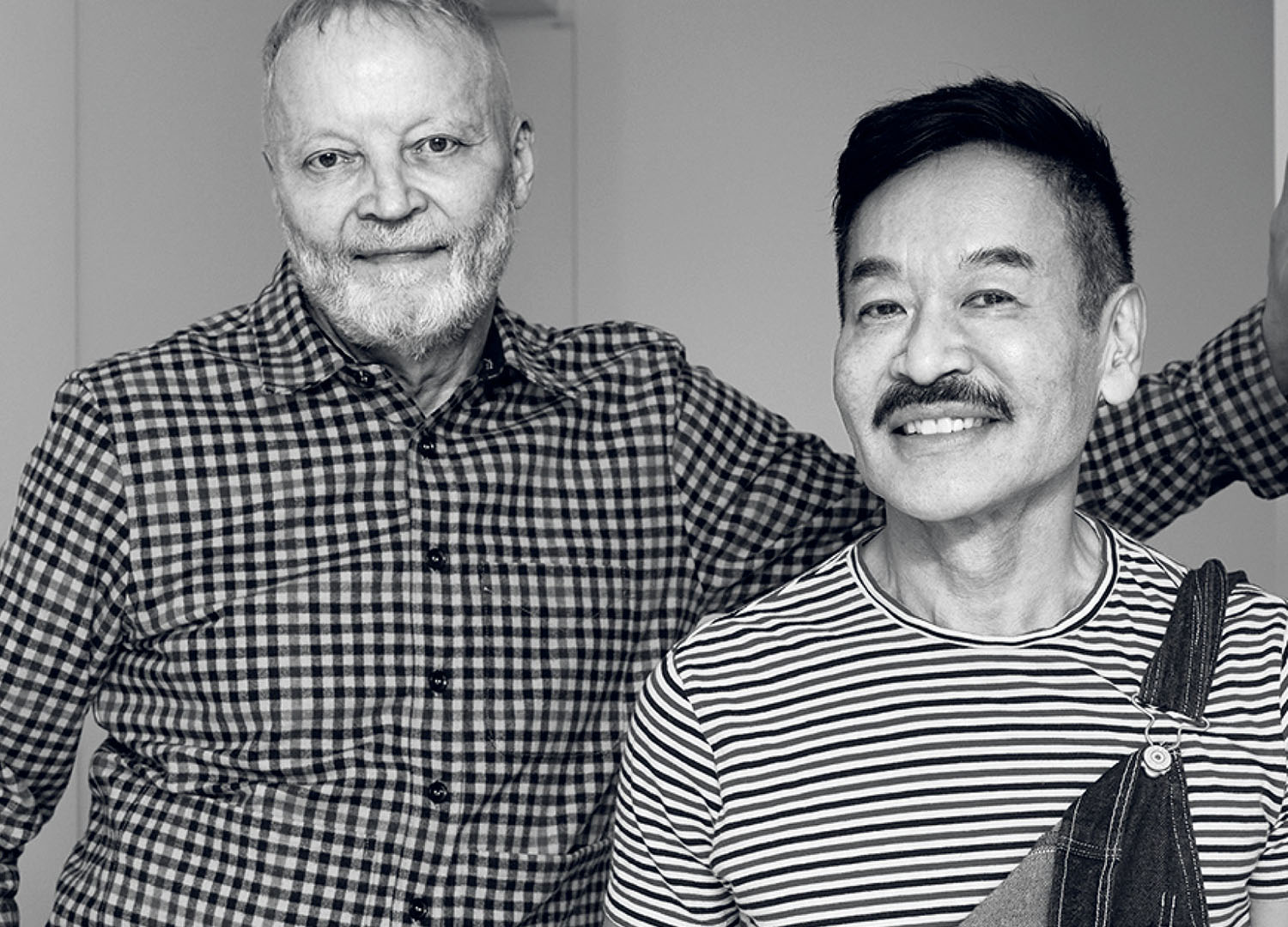 Two men smiling side-by-side, one wearing a plaid button-down shirt and the other wearing a striped t-shirt with overalls.