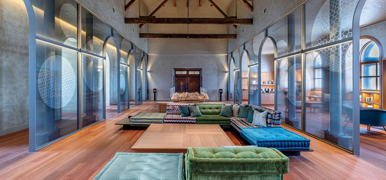 A large room with wood floors and exposed ceiling beams, featuring modular green and blue sofas arranged around a low wooden table, enclosed by tall arched glass partitions.